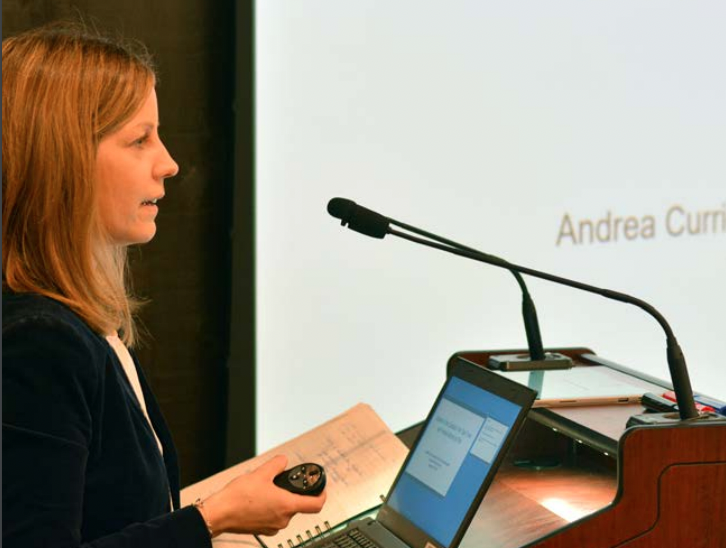 Andrea Currie, standing at a podium with a laptop visible on it, presenting at the Canadian Pain Care Forum meeting.