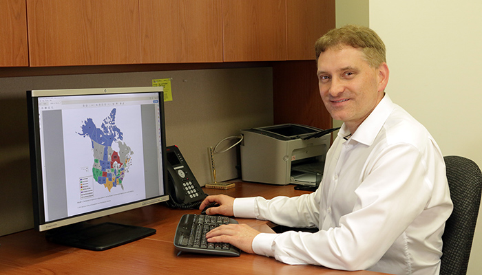 Jason Busse sitting at a desk in front of a computer screen that has a picture of Canada and the US on it.