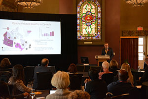 Jason Busse stands in behind a podium, next to a screen, presenting at the Chronic Post Surgical Pain Symposium.